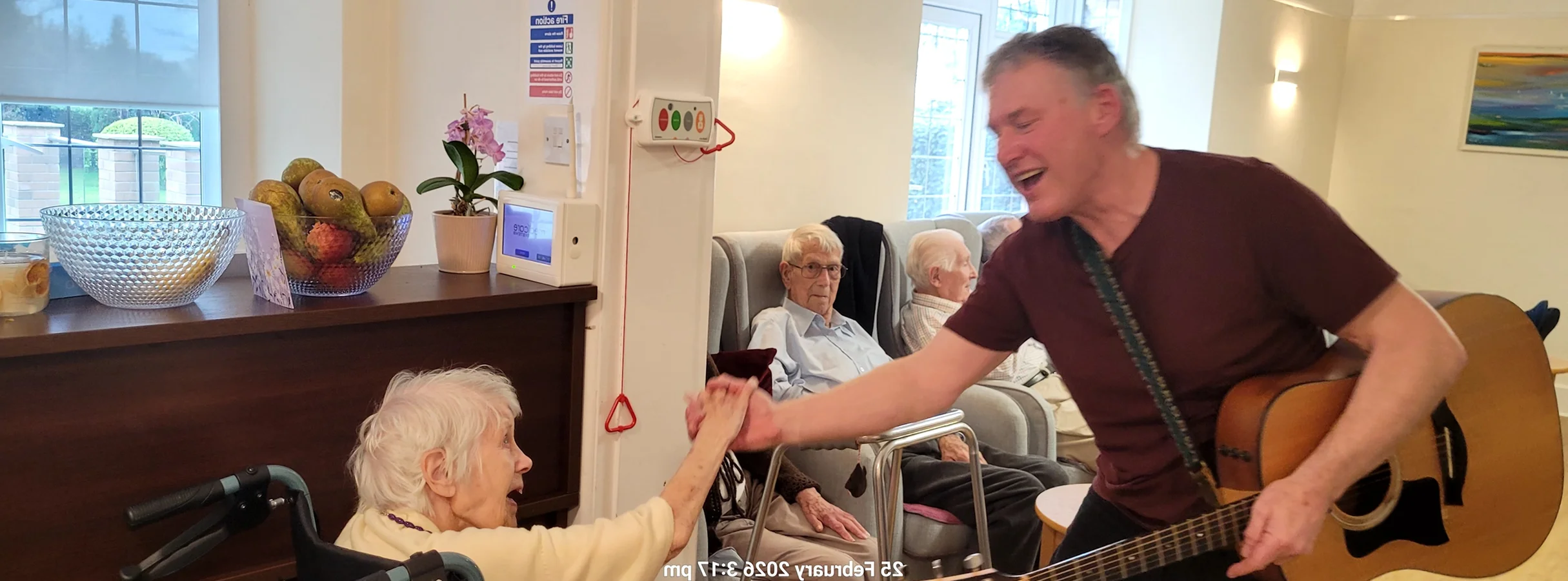Musician performing for seniors in a care home, playing guitar and engaging with an elderly woman in a wheelchair.