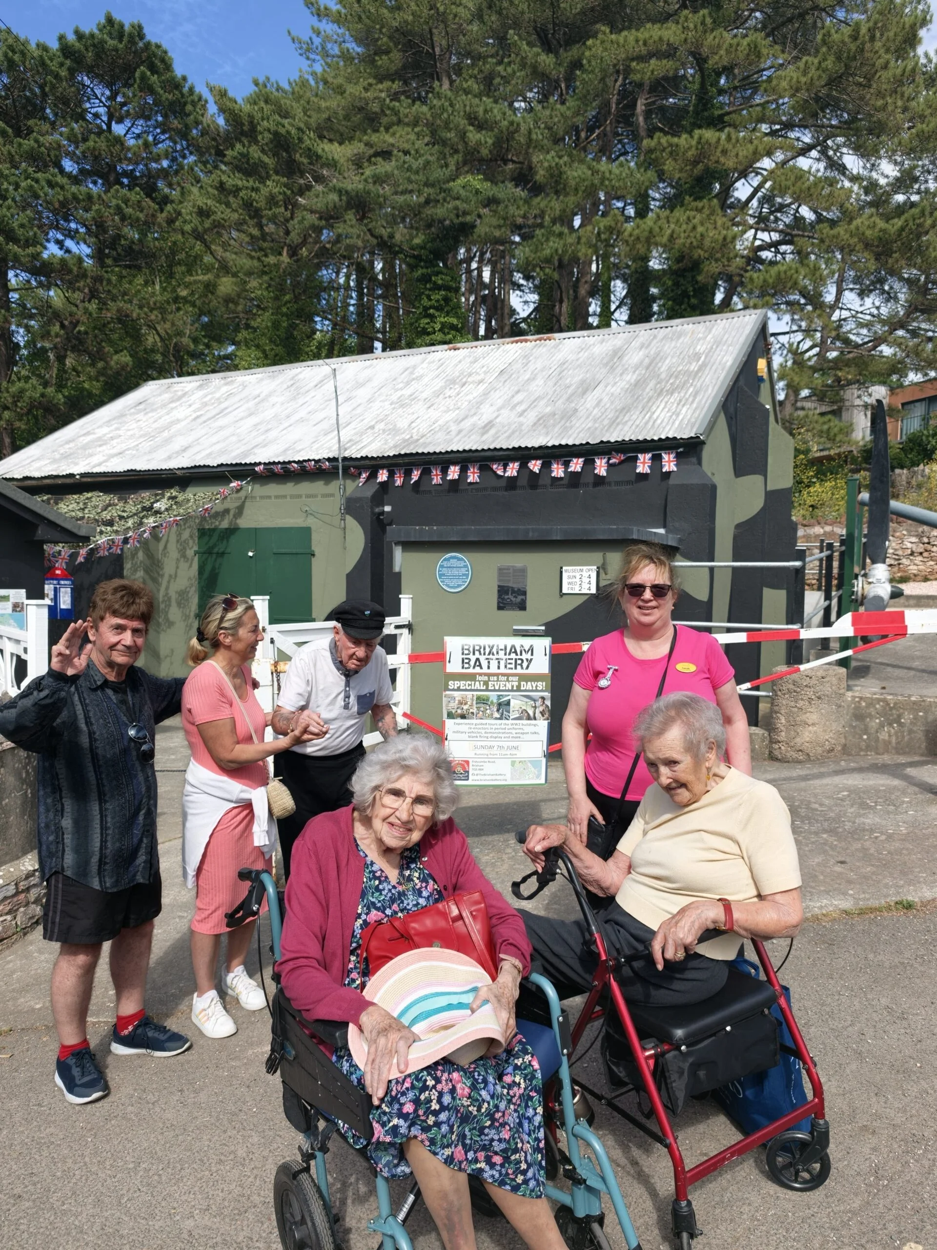 Group visiting Brixham Battery museum on a sunny day, two older women in wheelchairs, others smiling and standing nearby.