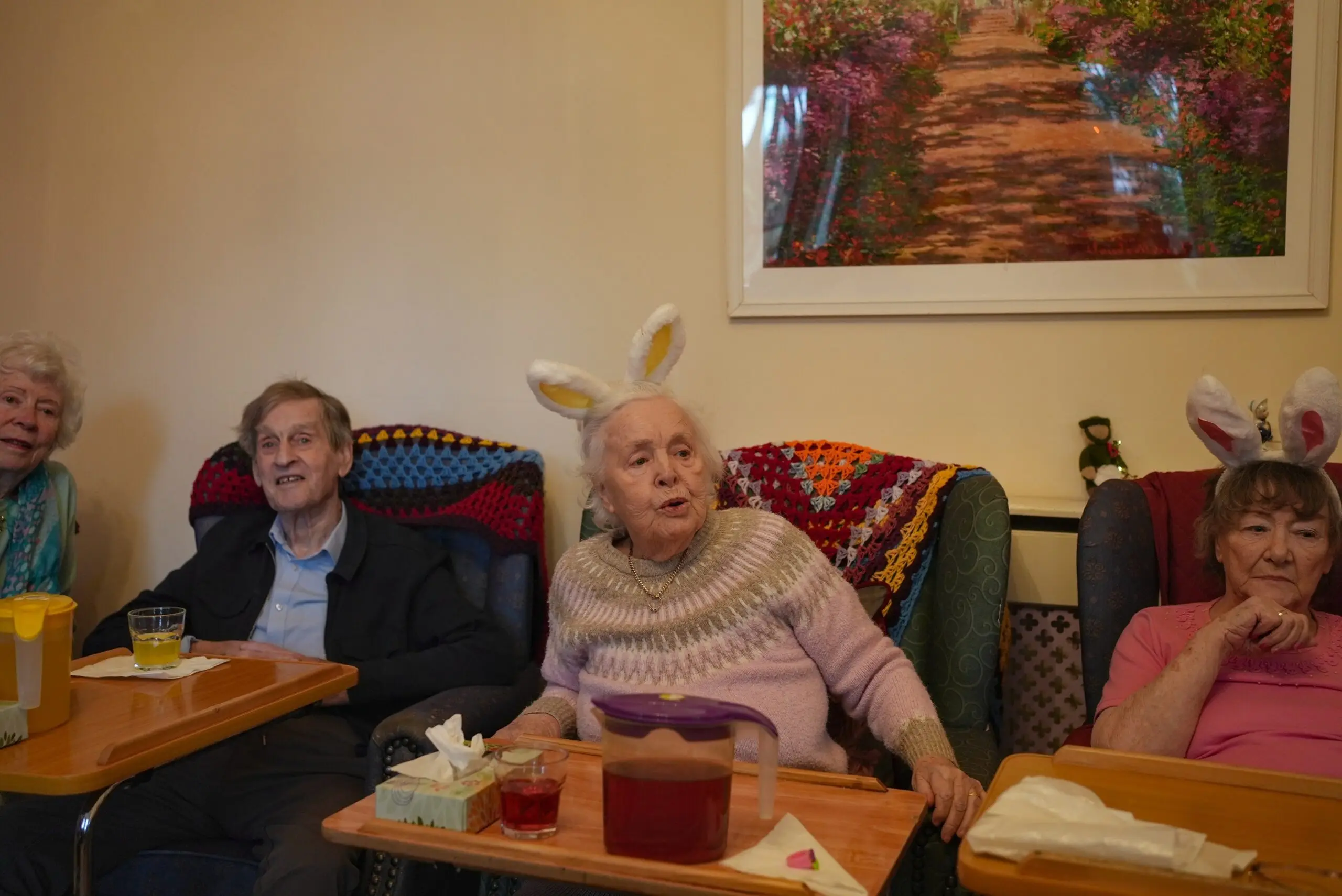 Elderly group enjoying time at a residential home, wearing bunny ear headbands, seated with drinks and colorful blankets.