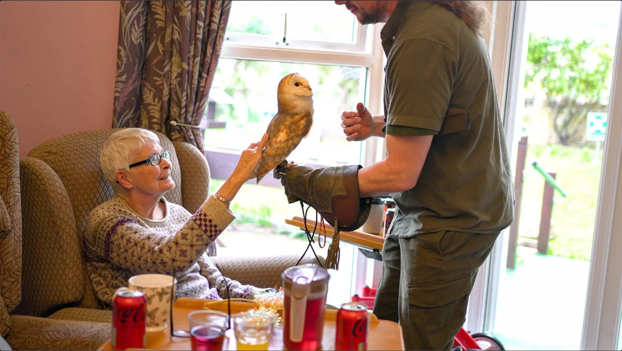 Elderly woman interacting with a barn owl on a man's glove in a cozy room.