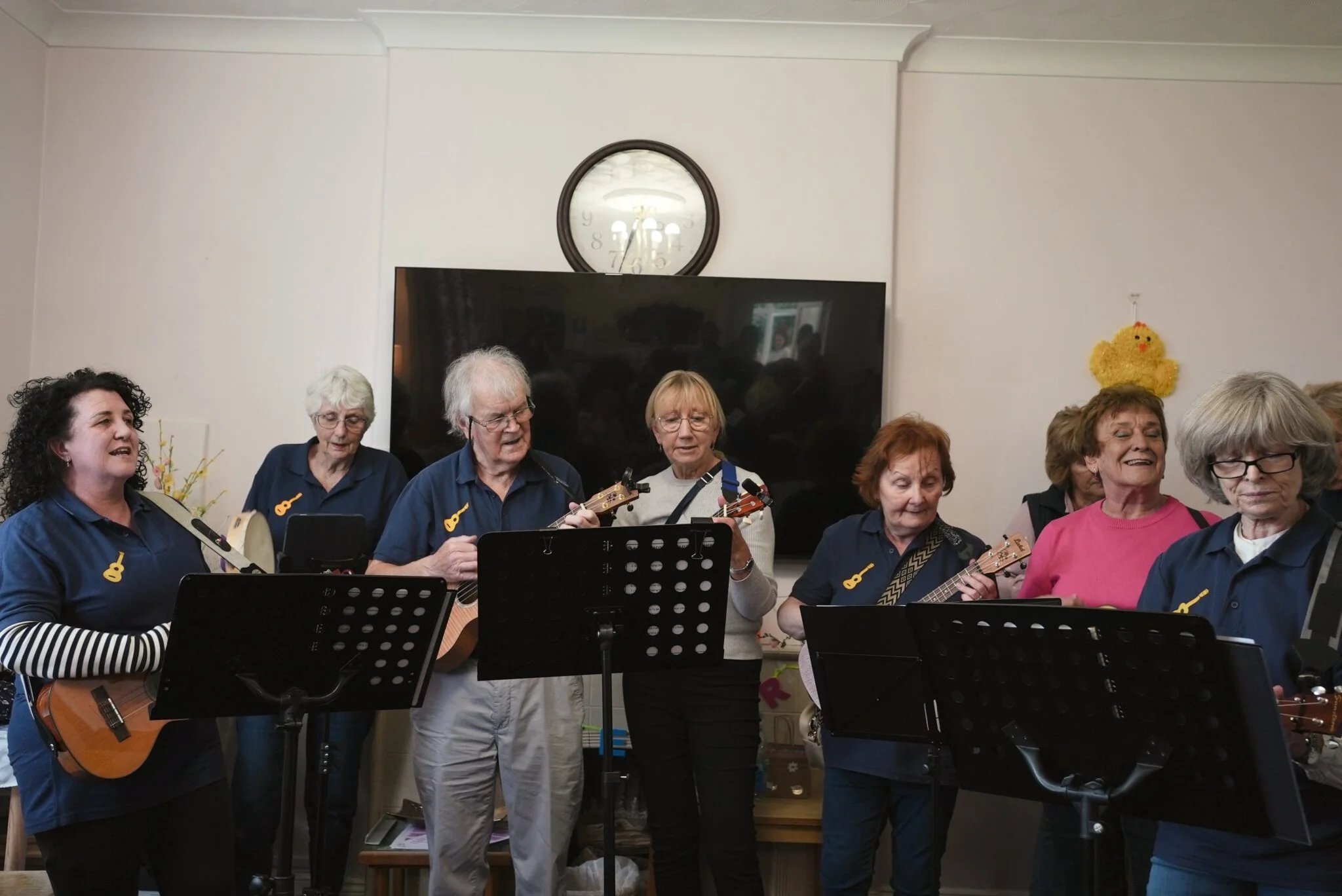 Senior adult ukulele group performing in a living room, with music stands and a wall clock in the background.