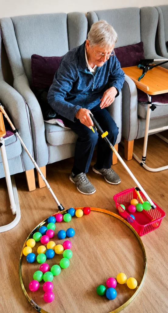 Elderly person using grabbers to collect colorful balls into a basket, engaging in a fun activity on the floor.
