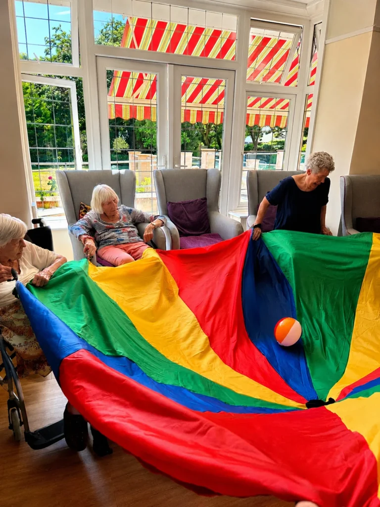 Seniors playing with a colorful parachute and beach ball in a bright room with large windows.