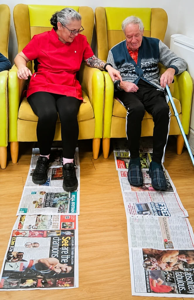 A caregiver holds hands with an elderly man in a care home, both seated with newspapers on the floor.
